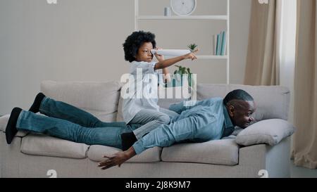 Joyeux famille afro-américaine père mûr se trouve sur le canapé dans la chambre joue avec petite fille mignon enfant fille assis sur le dos de papa regarde à travers Banque D'Images