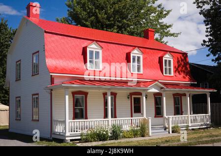 Ancienne maison de style cottage Canadiana vers 1825 avec revêtement en bois blanc et toit en mansarde en tôle rouge en été. Banque D'Images