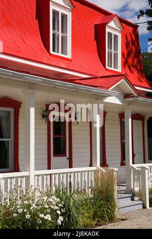Ancienne maison de style cottage Canadiana vers 1825 avec revêtement en bois blanc et toit en mansarde en tôle rouge en été. Banque D'Images