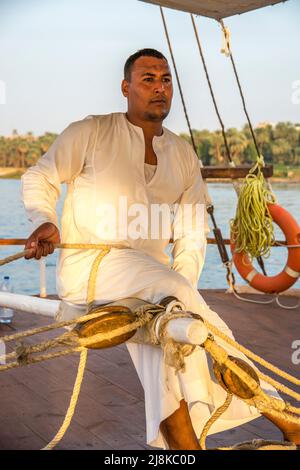 Membre de l'équipage tailler les voiles vêtues de vêtements traditionnels sur un bateau Dahabiya du Nil avec la rive du fleuve en arrière-plan Banque D'Images