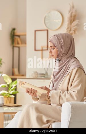Femme musulmane moderne portant le hijab rose pâle et des vêtements décontractés assis dans un fauteuil lisant un livre intéressant Banque D'Images