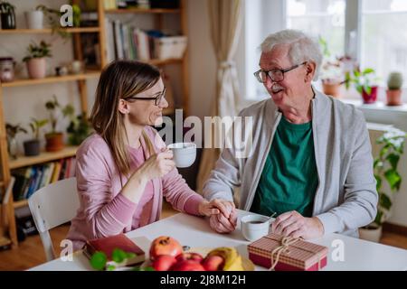 Fille adulte rendant visite à son père senior à la maison et prenant un café ensemble, en touchant la main. Banque D'Images