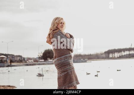 Bonne belle femme blonde en vêtements chauds marchant le long de la rive de la rivière seule pendant une journée fraîche et ensoleillée, se détendre et profiter, canards et cygnes nageant dans l'eau Banque D'Images