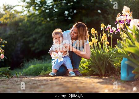 Bonne journée pour les enfants. Une jeune maman heureuse embrasse son bébé souriant tenant une fleur d'iris. En arrière-plan est un jardin avec des fleurs. L'été. Banque D'Images