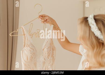 Élégante femme tenue de mariage robe blanche avec motif brodé sur le cintre dans la chambre. Portrait gros plan de Happy bride vue latérale, arrière-plan beige. Épouses Banque D'Images