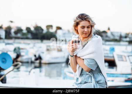 Femme blonde avec chemise autour du cou, tenant de savoureux verres de vin en vacances en voyage. Port avec bateaux. Copier l'espace Banque D'Images