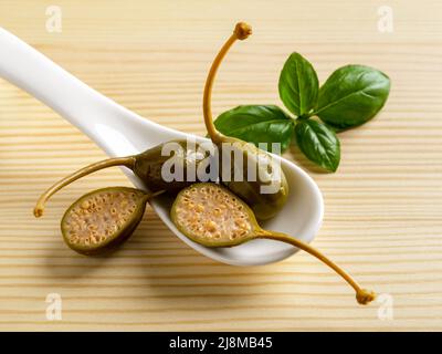 Baies de câpres marinées dans une cuillère blanche et feuille de basilic sur fond de bois. Câpres marinées entières et coupées en deux. Cuisine méditerranéenne. Banque D'Images