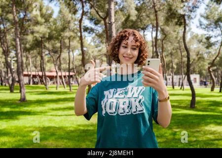 REDHEAD femme pointant au téléphone avec de grands sourires en se tenant sur un parc de la ville. Femme portant un t-shirt vert et une montre de poignet dorée. Femme sportive utilisant le ph Banque D'Images