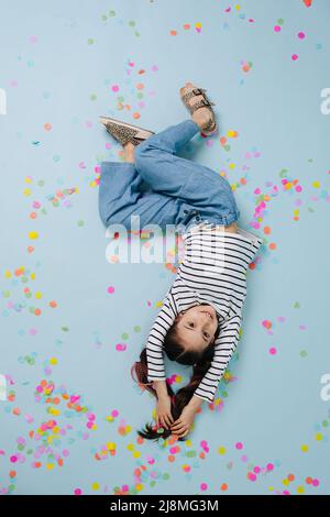 Image à l'envers d'une jolie fille souriante, couchée sur un sol bleu. Entouré de confettis colorés. Banque D'Images