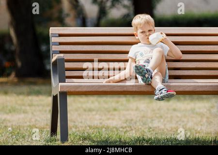 Un petit garçon avec est assis sur le banc dans le parc et boit le jus de la tasse en plastique. Banque D'Images