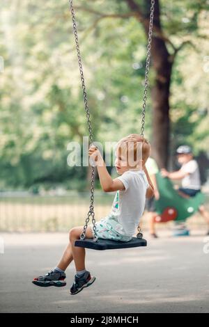 Joyeux petit garçon balançant sur la chaîne balançoire à l'aire de jeux. Banque D'Images