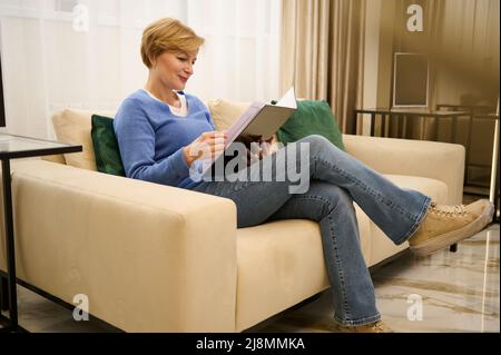 Une charmante femme de race blanche d'âge moyen est assise sur un canapé confortable dans le hall d'un salon de beauté et se jette dans un magazine tout en attendant un Banque D'Images