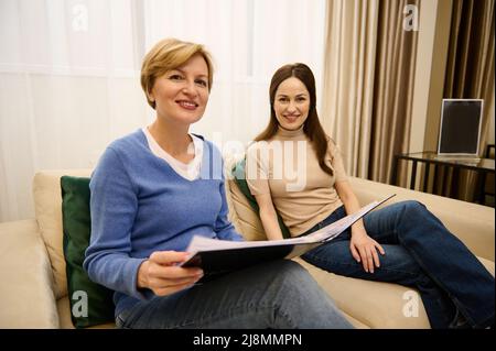 Deux agréables femmes européennes d'âge moyen avec un catalogue, sourire regardant la caméra, s'asseoir sur un canapé confortable dans un intérieur moderne et confortable d'un bureau Banque D'Images