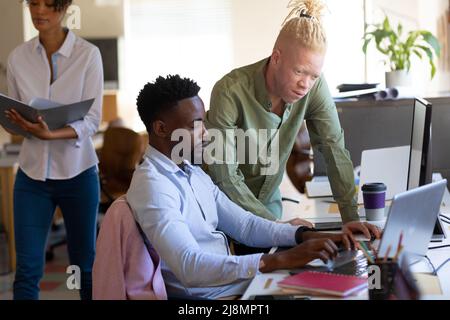 Albino homme d'affaires de taille moyenne assistant un collègue de sexe masculin utilisant un ordinateur portable au bureau Banque D'Images