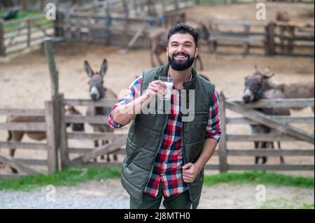 Homme barbu aux cheveux foncés en chemise à carreaux avec un verre et du lait près du bétail Banque D'Images