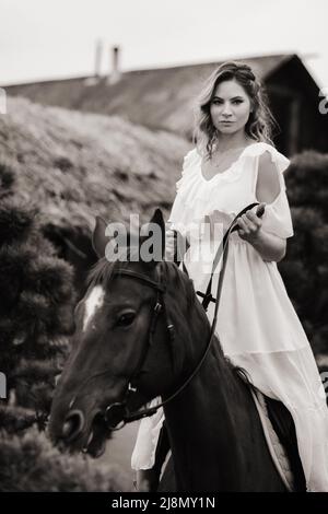 Une femme dans une sundress blanche à cheval près d'une ferme. Photo en noir et blanc Banque D'Images