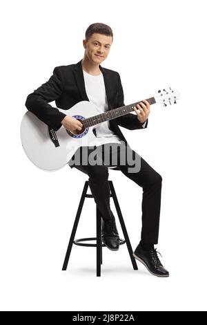 Jeune homme élégant en costume noir jouant une guitare acoustique assise sur une chaise isolée sur fond blanc Banque D'Images