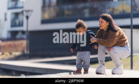 Printemps. Jeune mère brune adulte tenant son petit fils multiracial sur la chaussée. Famille profitant du temps ensoleillé. Photo de haute qualité Banque D'Images