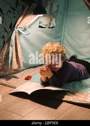 un garçon aux cheveux bouclés examine un livre dans un wigwam Banque D'Images