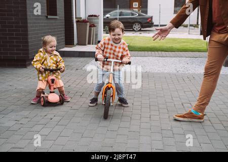 Enfants jouant à la course de vélo et de scooter dans la cour de la maison. Sœur et frère à cheval rapide autour de la maison privée. Concours de frères et sœurs de famille. Enfants Banque D'Images