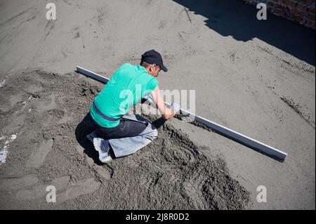 Vue de dessus du travailleur masculin plaçant le rail de table sur le sol recouvert de mélange sable-ciment sur le chantier de construction.Homme nivelant la surface avec bord droit tout en criant le sol à l'extérieur dans le nouveau bâtiment. Banque D'Images