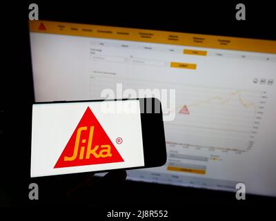 Personne tenant un téléphone portable avec le logo de la société suisse de produits chimiques spécialisés Sika AG sur écran en face de la page web d'affaires. Mise au point sur l'affichage du téléphone. Banque D'Images