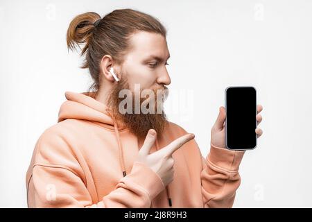 Scandinave beau homme sérieux tenant un téléphone et pointe vers l'écran noir maquette avec casque isolé sur fond gris. Un homme calme avec du gingembre Banque D'Images