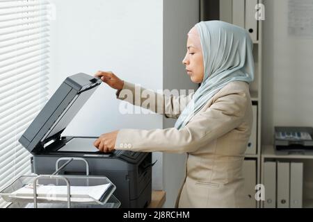 Vue latérale d'une jeune femme musulmane adulte moderne portant le hijab travaillant au bureau pour faire des copies ou numériser des documents Banque D'Images