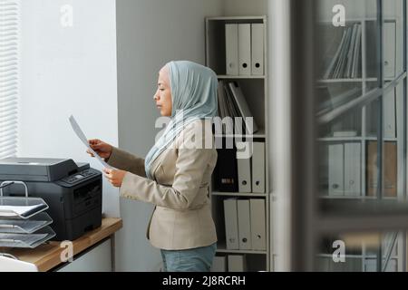 Vue latérale d'une femme musulmane moderne portant le hijab travaillant dans un bureau debout devant l'imprimante vérifiant des copies de document Banque D'Images
