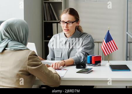Un agent consulaire caucasien moderne assis à son bureau devant une femme musulmane lui expliquant comment remplir le formulaire de demande Banque D'Images