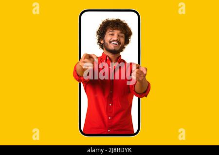 Beau mec indien mauriquement regardant depuis un immense smartphone et pointe les doigts vers l'appareil photo, l'homme latin vous choisissant, offrant de rejoindre un jeu en ligne ou une communauté virtuelle, studio tourné isolé sur jaune Banque D'Images