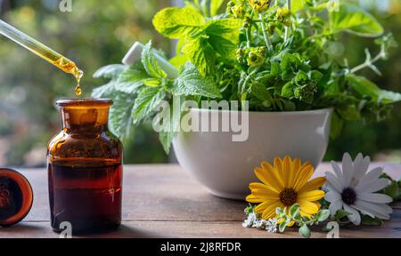 Médecine alternative de santé. Goutte d'huile essentielle tombant dans une bouteille en verre, gros plan. Cosmétiques naturels, sérum et vitamines. Herbes fraîches dans un mortier sur Banque D'Images