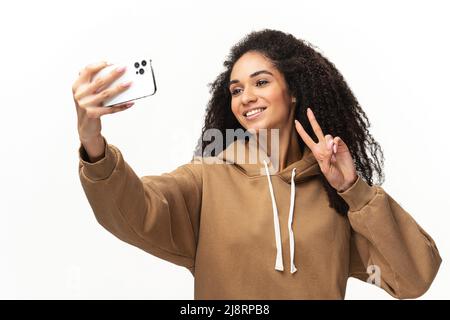 Une jeune femme afro-américaine insouciante, aux cheveux bouclés, qui prend le selfie sur son smartphone, enregistre des messages vidéo, diffuse sur des réseaux sociaux, isolée sur du blanc Banque D'Images