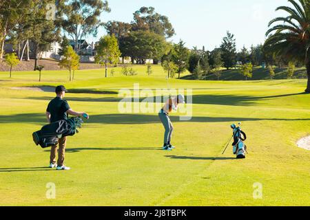Vue arrière d'un jeune homme caucasien debout avec un sac de golf tandis qu'un ami afro-américain joue au golf Banque D'Images