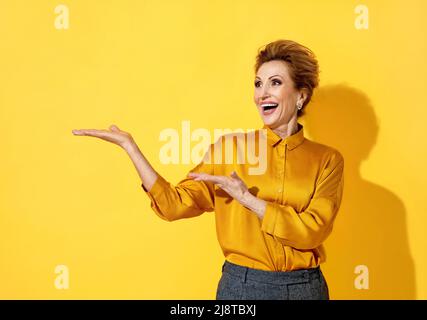 Femme joyeuse montrant un espace de copie vide (publicités, promotion, recommander). Photo d'une femme âgée attrayante en chemise jaune sur fond jaune Banque D'Images