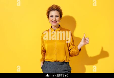 Bonne femme âgée montre le pouce vers le haut, comme un geste. Photo de la femme en chemise jaune sur fond jaune. Émotions et sentiments agréables concept. Banque D'Images
