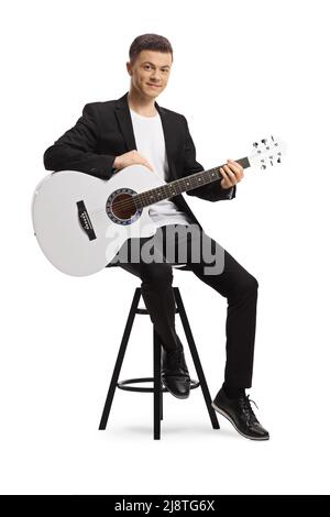 Jeune homme élégant en costume noir assis sur une chaise et tenant une guitare acoustique blanche isolée sur fond blanc Banque D'Images