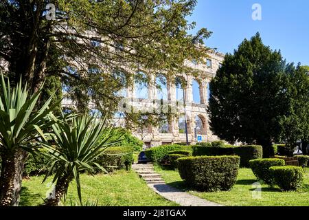 Parc avec pelouse bien entretenue, palmiers, oliviers, haies et cyprès devant les ruines d'un amphithéâtre romain Banque D'Images