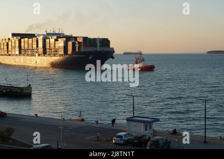 Immense navire à conteneurs entièrement chargé de CMA CGM compagnie entrant dans le port de Koper pendant le coucher du soleil avec le ciel orange. Le bateau est assisté par bateau à vapeur orange Banque D'Images