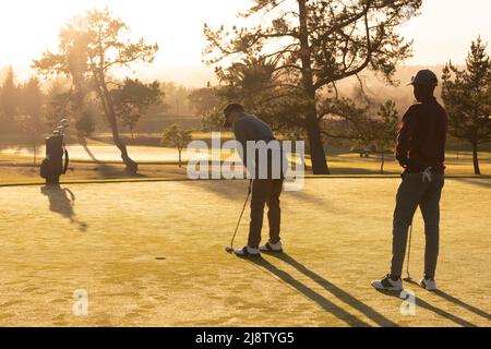 Jeunes amis multiraciaux jouant au golf sur un parcours de golf contre un ciel dégagé au coucher du soleil Banque D'Images
