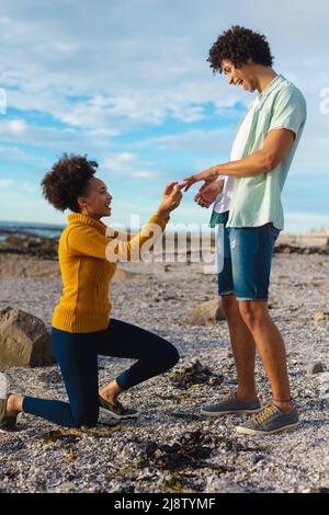 Bonne jeune femme afro-américaine proposant un copain afro-américain excité tout en s'agenouillant à la plage Banque D'Images