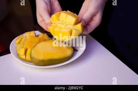 La femme coupe la mangue mûre fraîche en cubes pour que la pulpe de fruit se sépare facilement de la peau à l'aide d'un couteau de cuisine. Banque D'Images