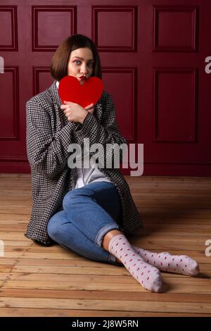 Femme à poil court en veste assise sur parquet et arrière-plan rouge tenant le cœur rouge devant son menton Banque D'Images