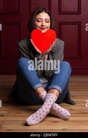 Femme souriante aux cheveux courts en veste assise sur un sol en bois jambes croisées et arrière-plan rouge tenant la forme du cœur rouge devant son menton Banque D'Images