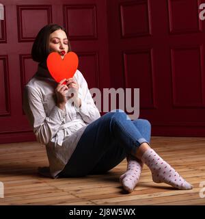 Femme aux cheveux courts avec les yeux fermés et les lèvres formant baiser assis sur le plancher de bois jambes croisées et arrière-plan rouge tenant la forme de coeur rouge devant Banque D'Images