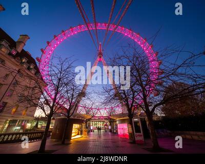 Le London Eye, rose illuminé, vu de la promenade de la Reine la nuit, avec deux silhouetés personnes au loin. Londres. Banque D'Images