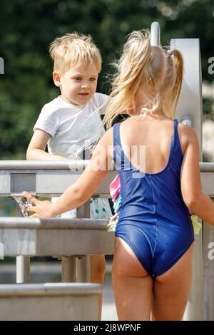 Un petit garçon heureux et une belle fille dans un maillot de bain bleu joue avec un robinet d'eau dans un parc de la ville. Équipement d'eau spécial pour les jeux pour enfants sur un summe chaud Banque D'Images