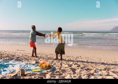 Couple afro-américain tenant les mains et dansant sur la plage de sable contre la mer et ciel clair en été Banque D'Images