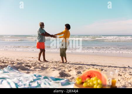 Couple afro-américain tenant les mains et dansant sur la plage de sable contre la mer et le ciel bleu en été Banque D'Images