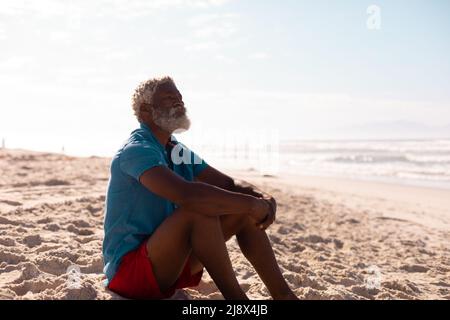 Homme senior afro-américain barbu avec des cheveux gris assis sur une plage de sable contre le ciel Banque D'Images